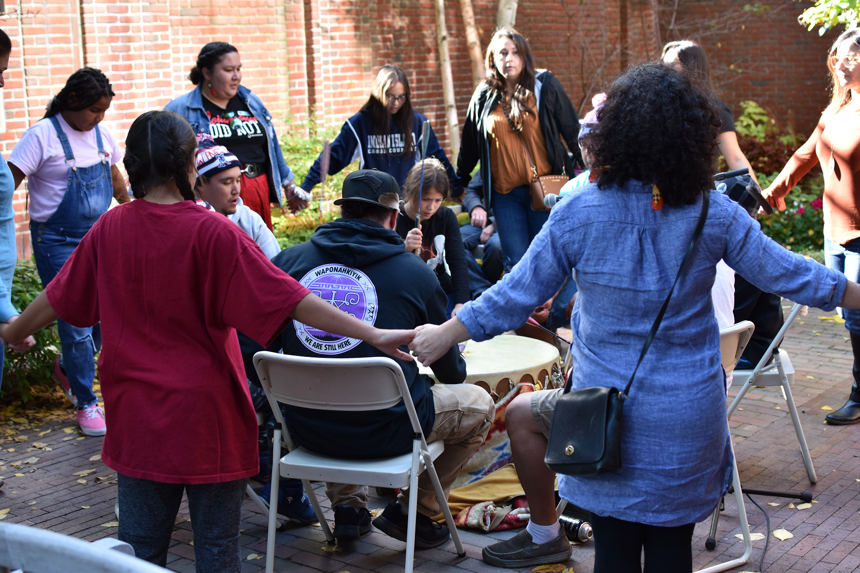 A group of Wabanaki tribal members stand, with hands joined, in a circle around a drum on Indigenous People's Day.