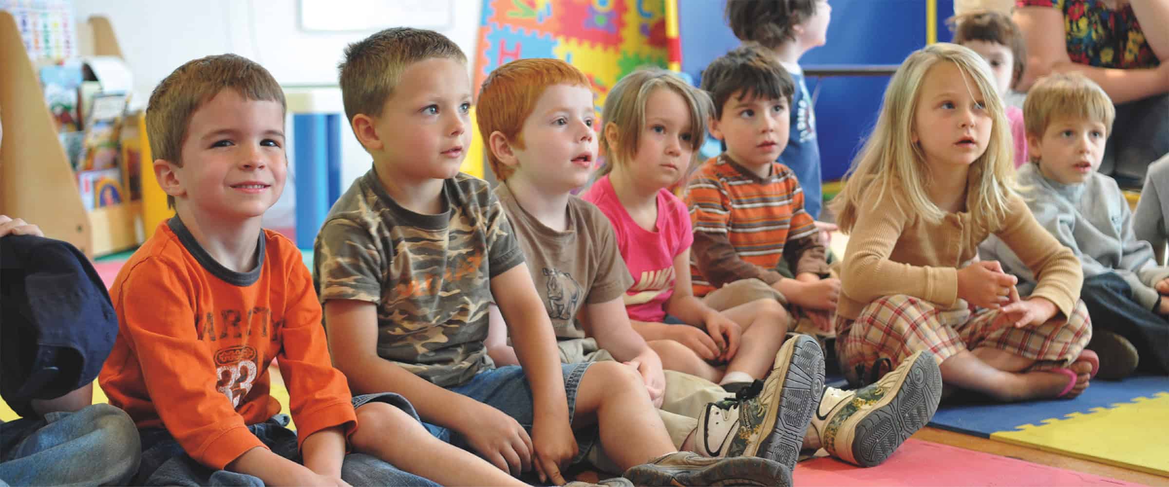 A row of young children sit cross-legged on colorful play mats at Coastal Kids Preschool in Damariscotta, Maine.