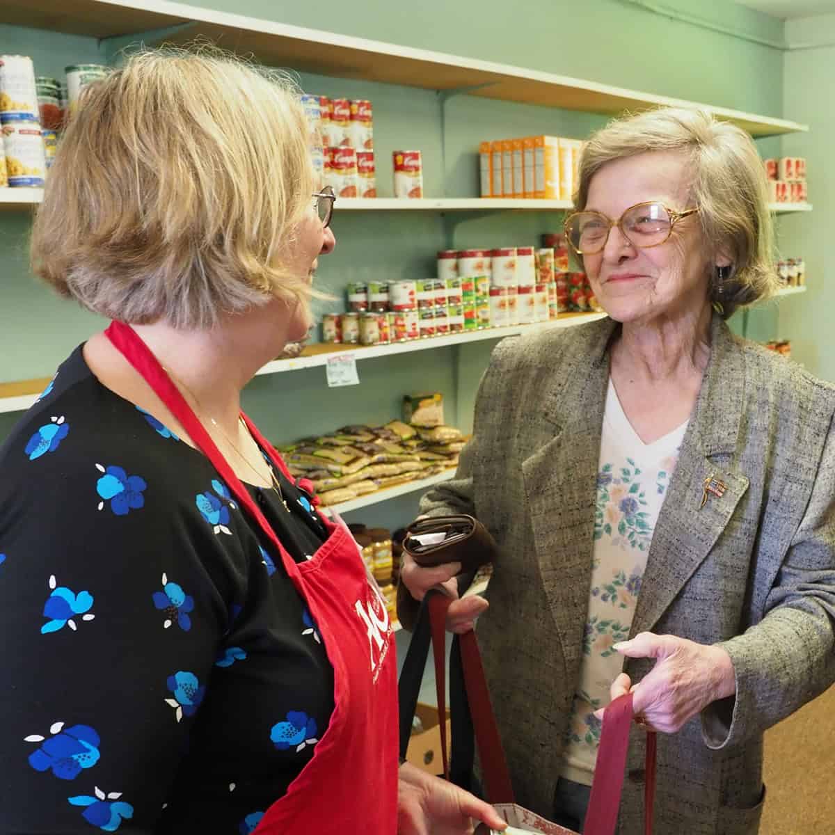 Two women talking in a store, one with a red apron, the other with glasses and a cane.