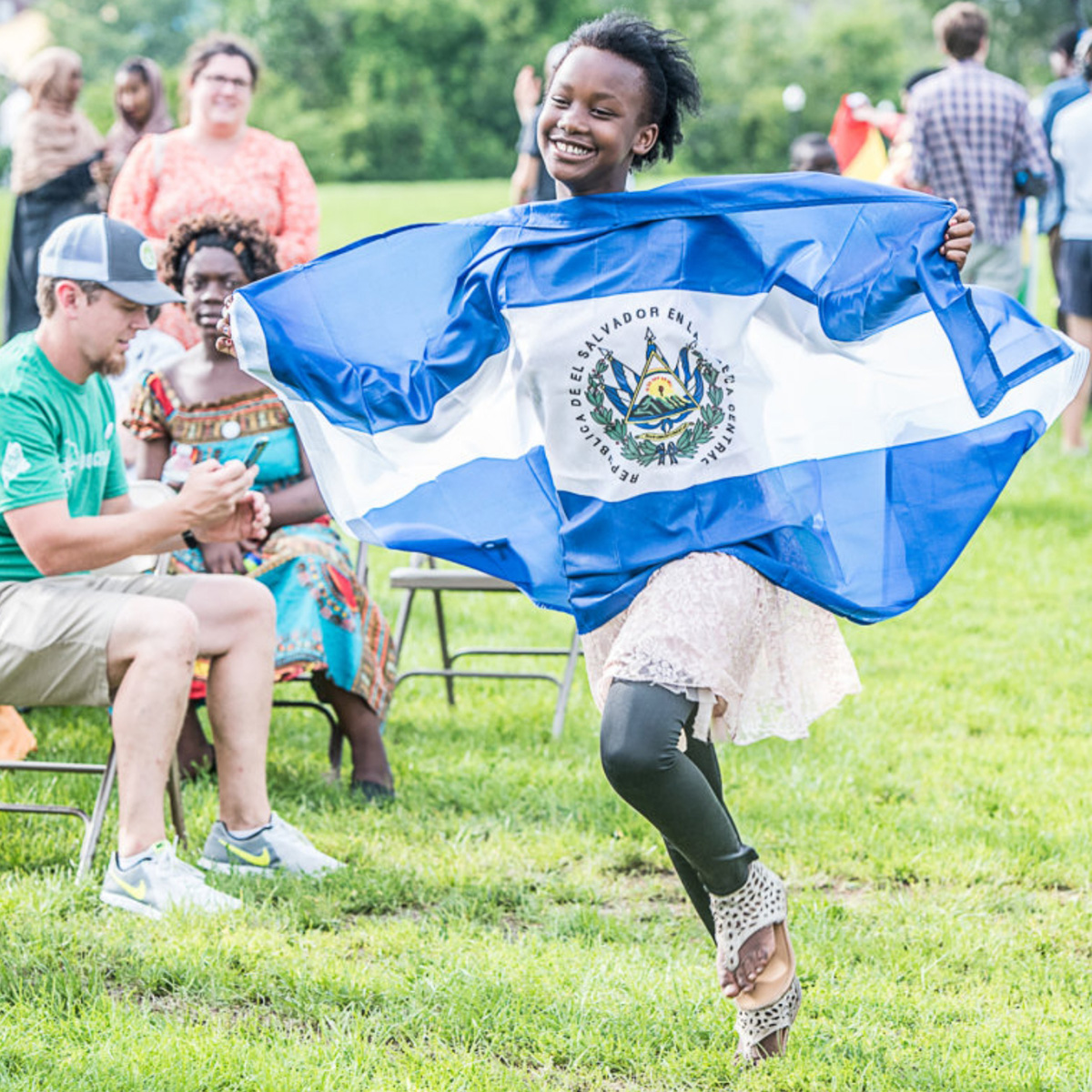 A smiling young girl holds a Republic of El Salvador flag as she runs in front of onlookers at a community celebration in Lewiston, Maine.