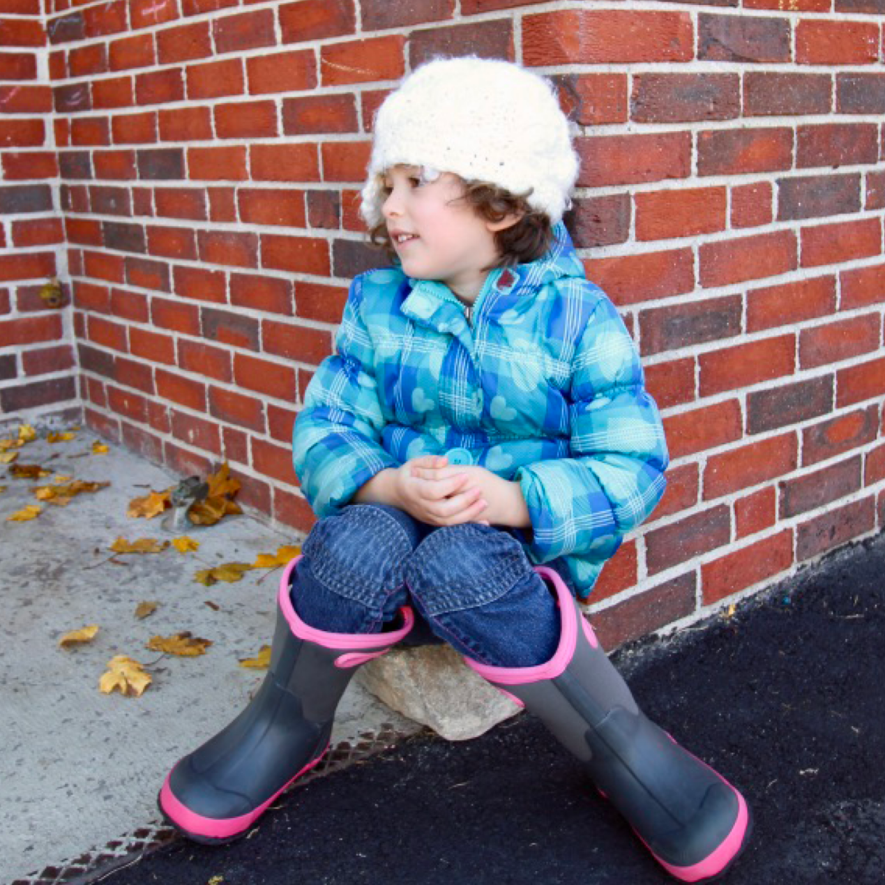 Girl sitting against brick building and wearing blue jacket, white hat, and black rain boots with pink trim