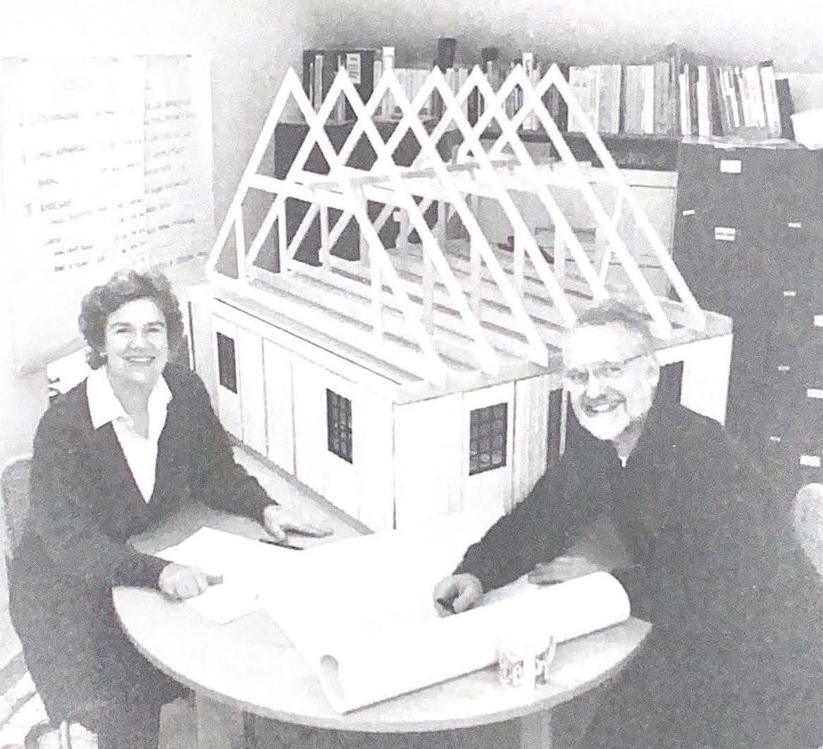 Black and white photo of man and woman sitting at table with blue prints and architectural model of a house