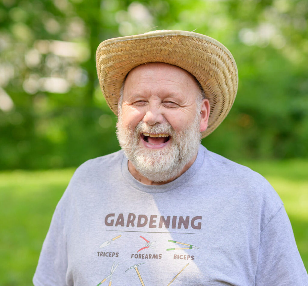 Older white man with white beard, who lives in Bath Housing. He is wearing straw hat and gray T-sirt that says GARDENING.