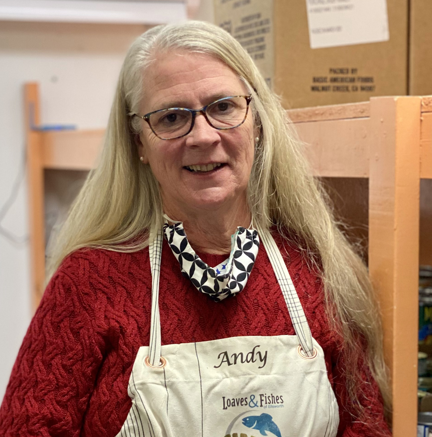 A food bank volunteer with an apron on smiles at the camera while standing in front of shelves of food and supplies for local families in need.