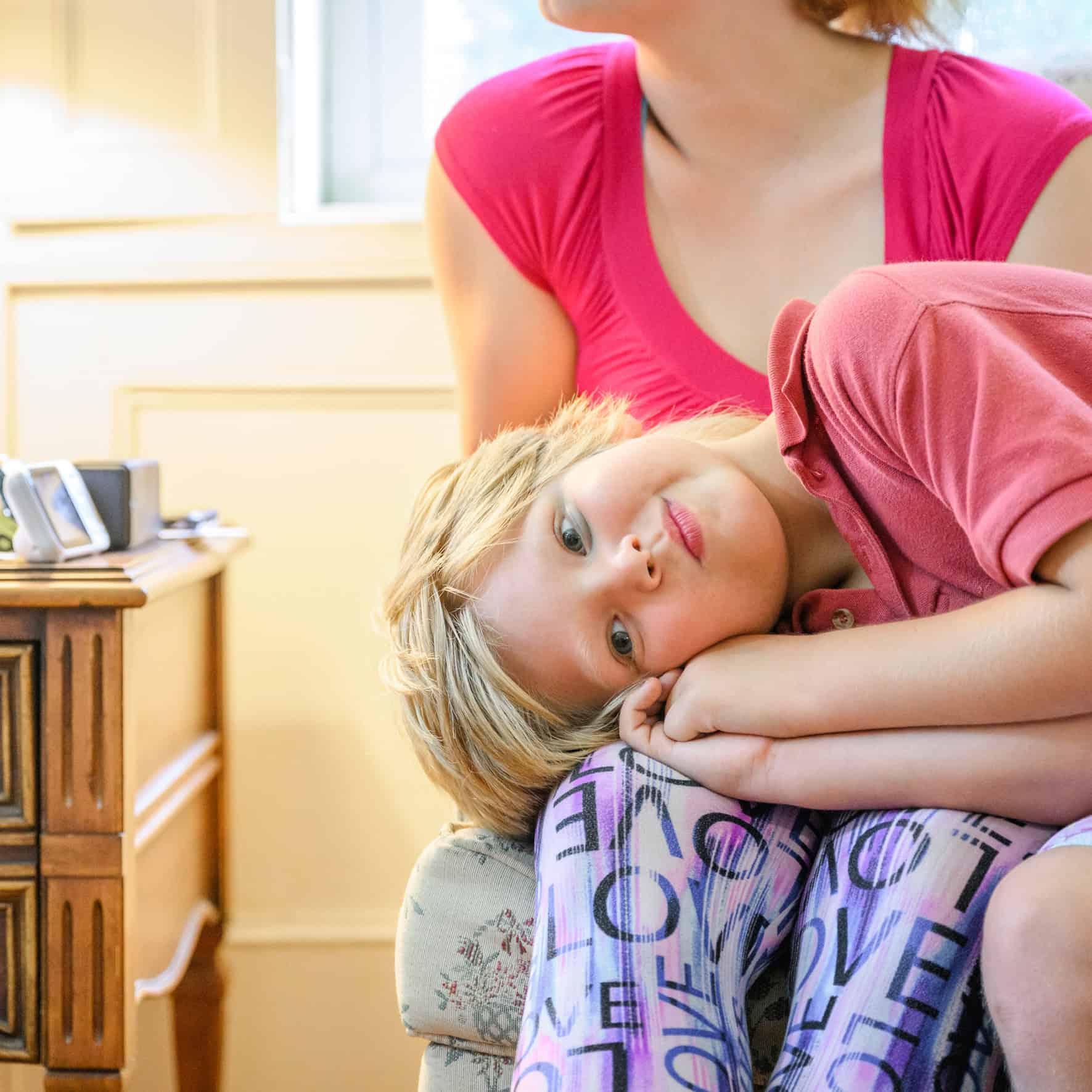 Young girl resting on a couch with a woman in a pink shirt nearby.
