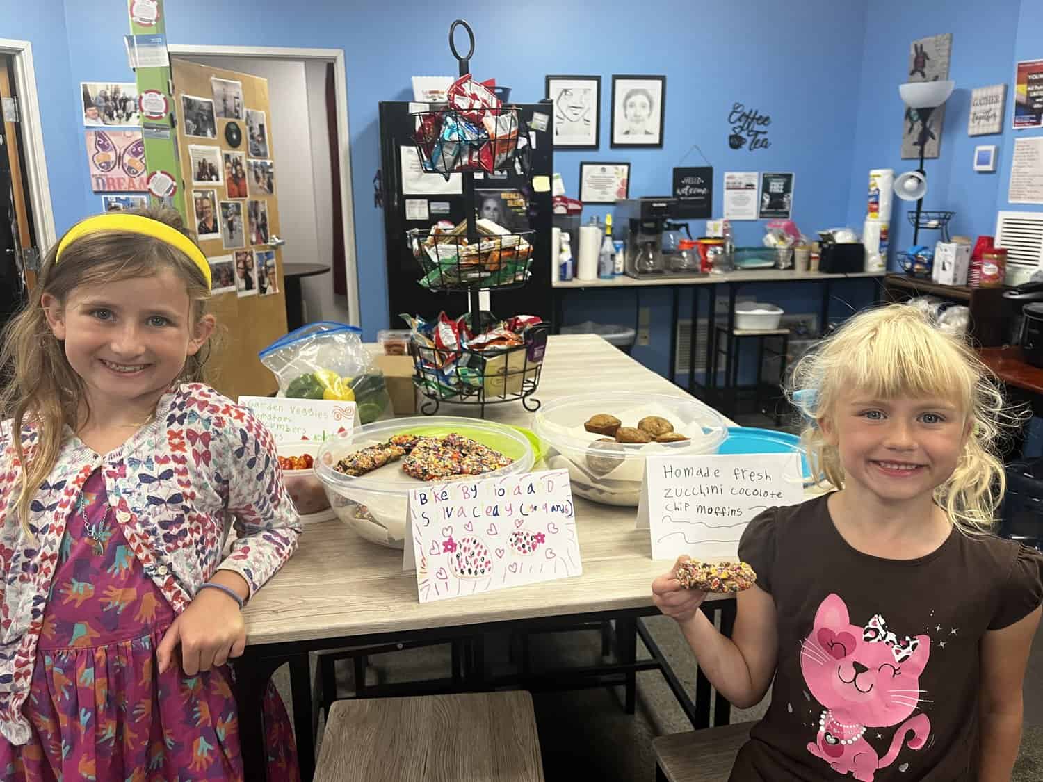 Two smiling young girls with baked goods at a community event table.