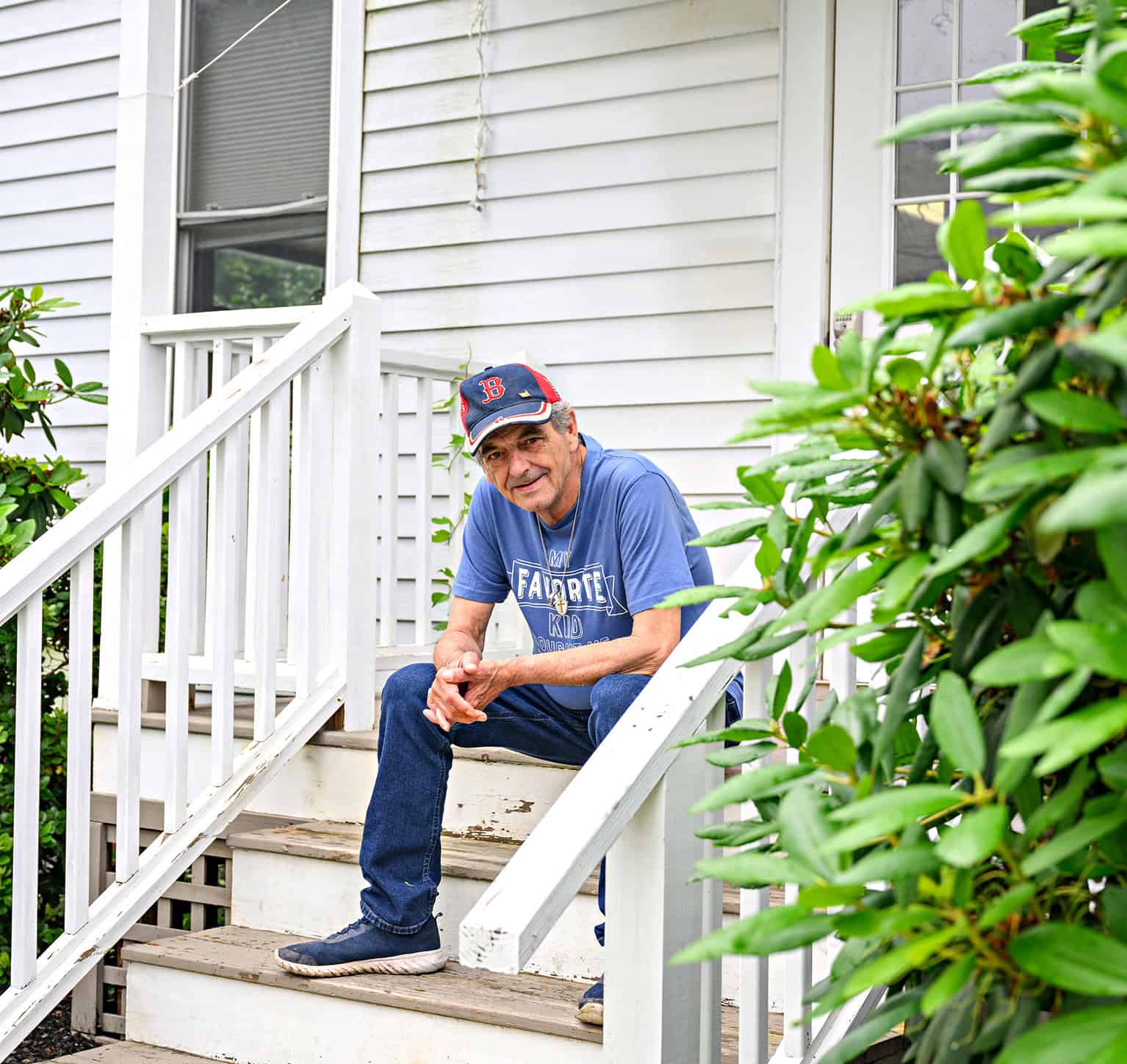 Man sitting on front porch steps of a house with greenery nearby.