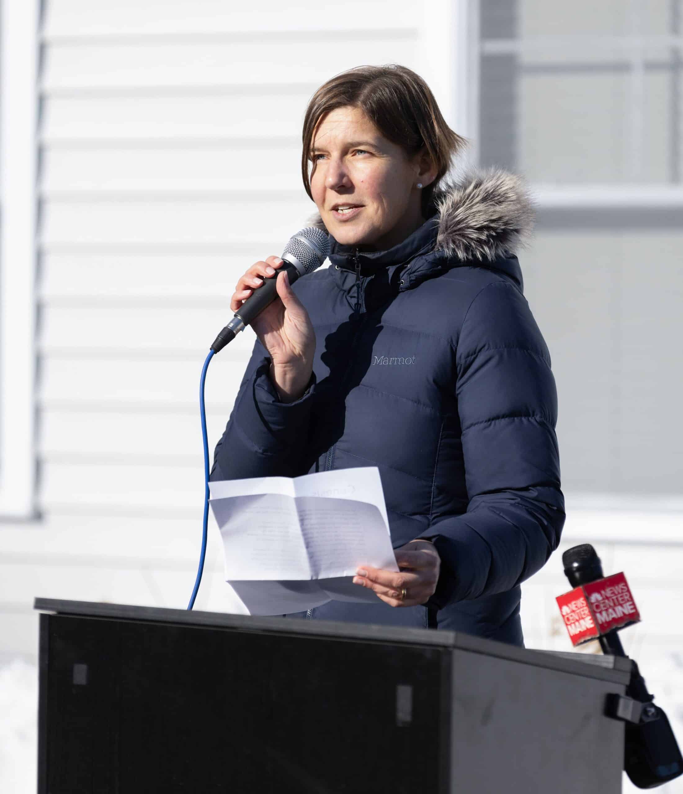 Woman speaking into microphone at outdoor event with white building background.