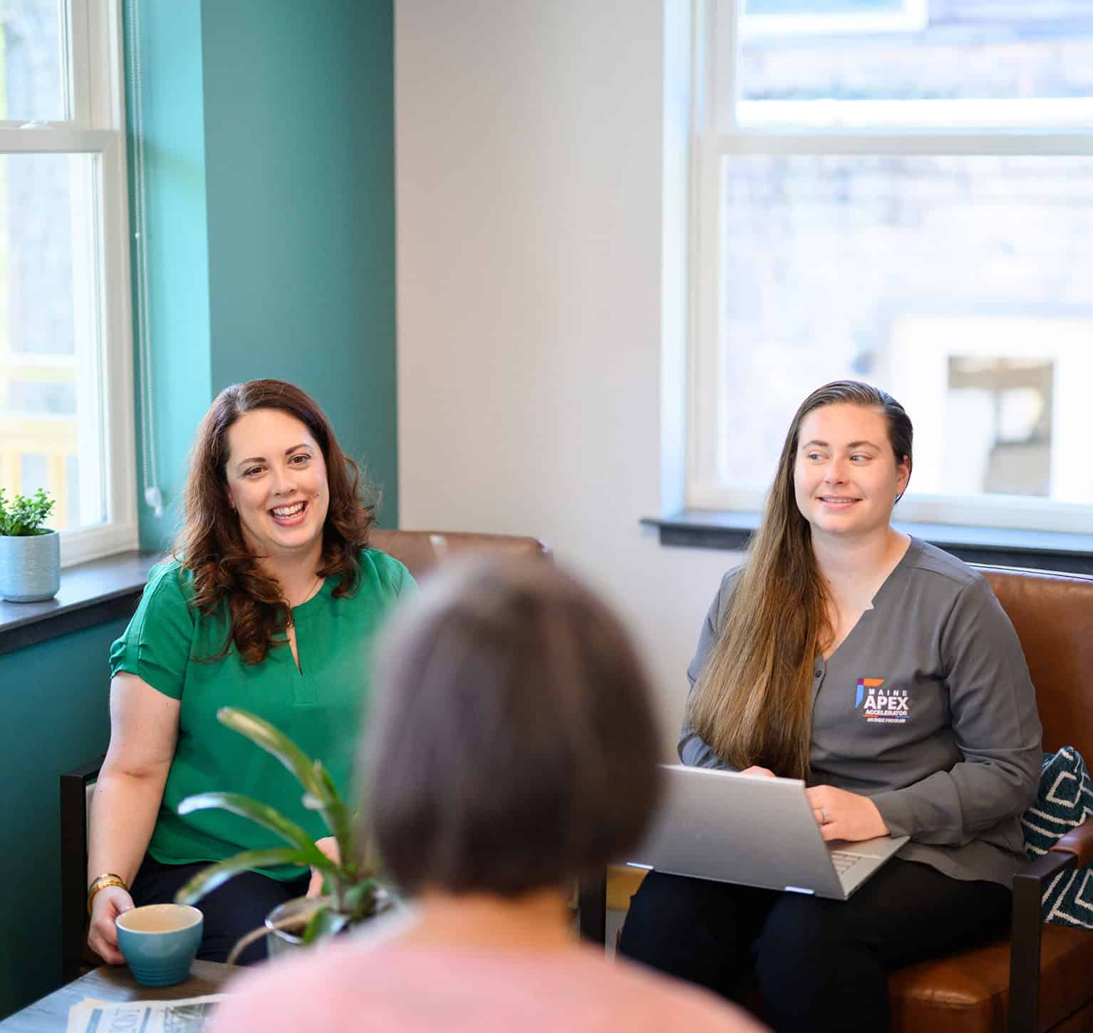Two women in a meeting room with a third person, smiling and engaging.