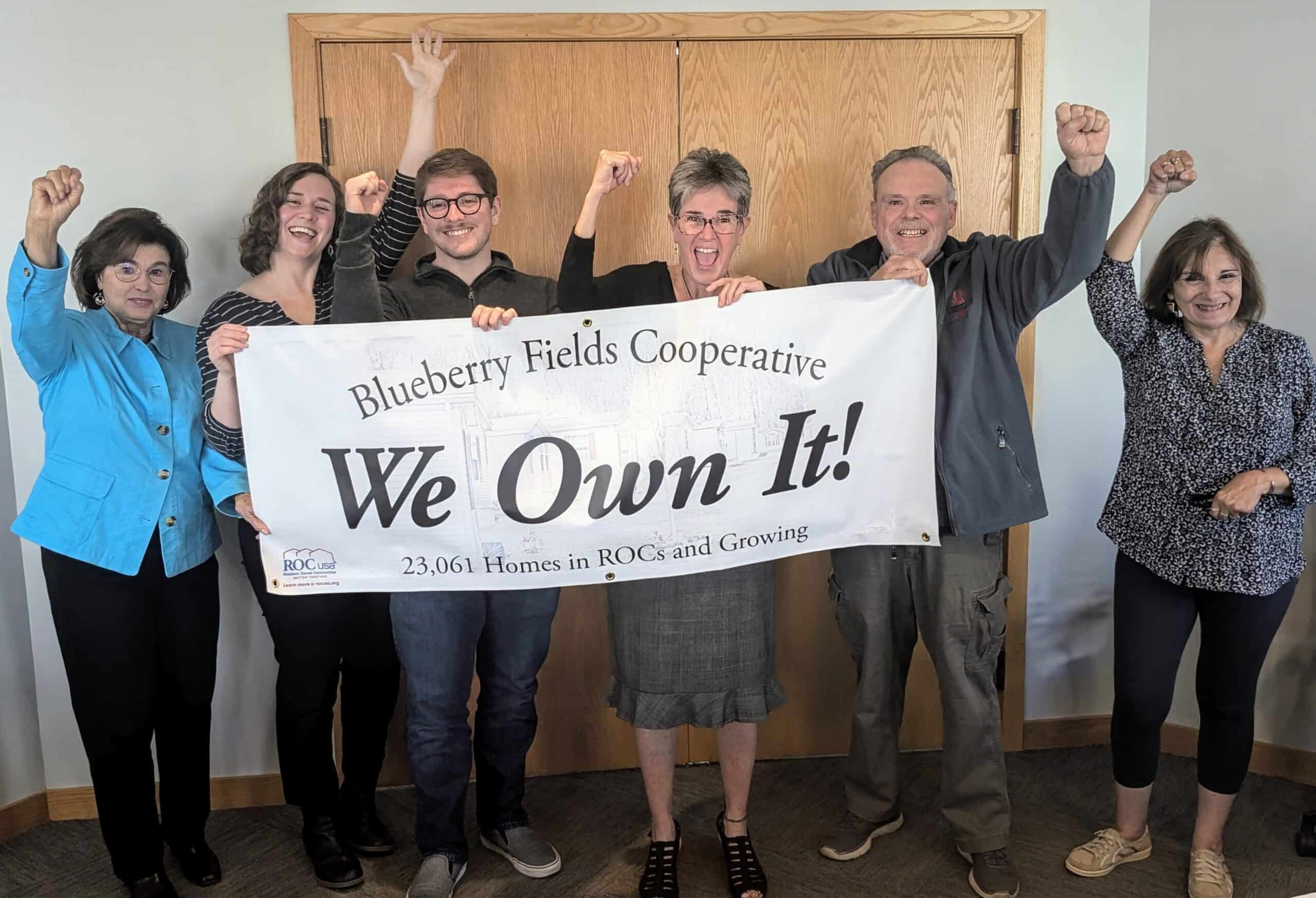 Group of diverse people holding a 'We Own It!' banner at Blueberry Fields Cooperative.