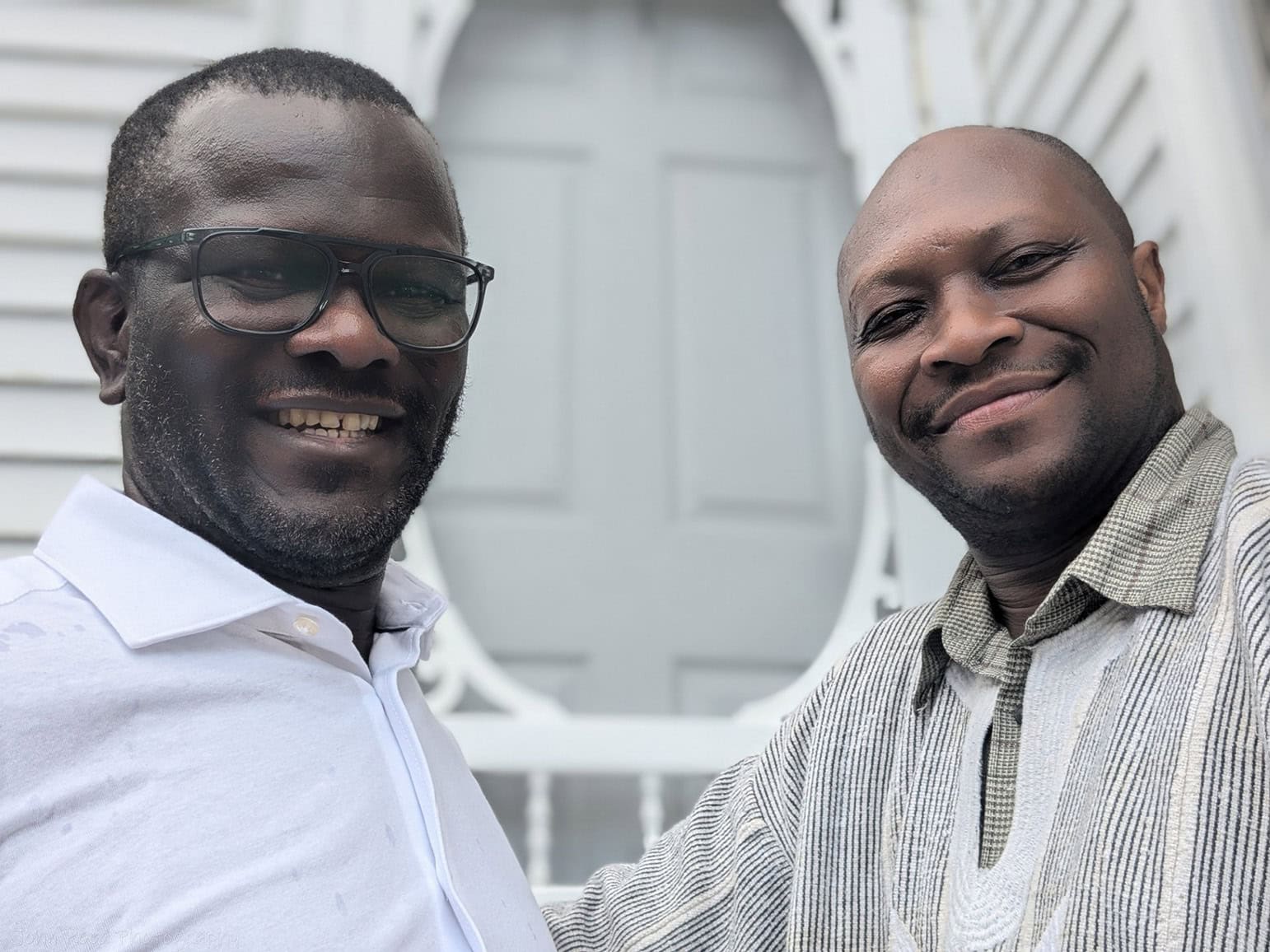Two men smiling outdoors, one wearing glasses, in front of a white structure.