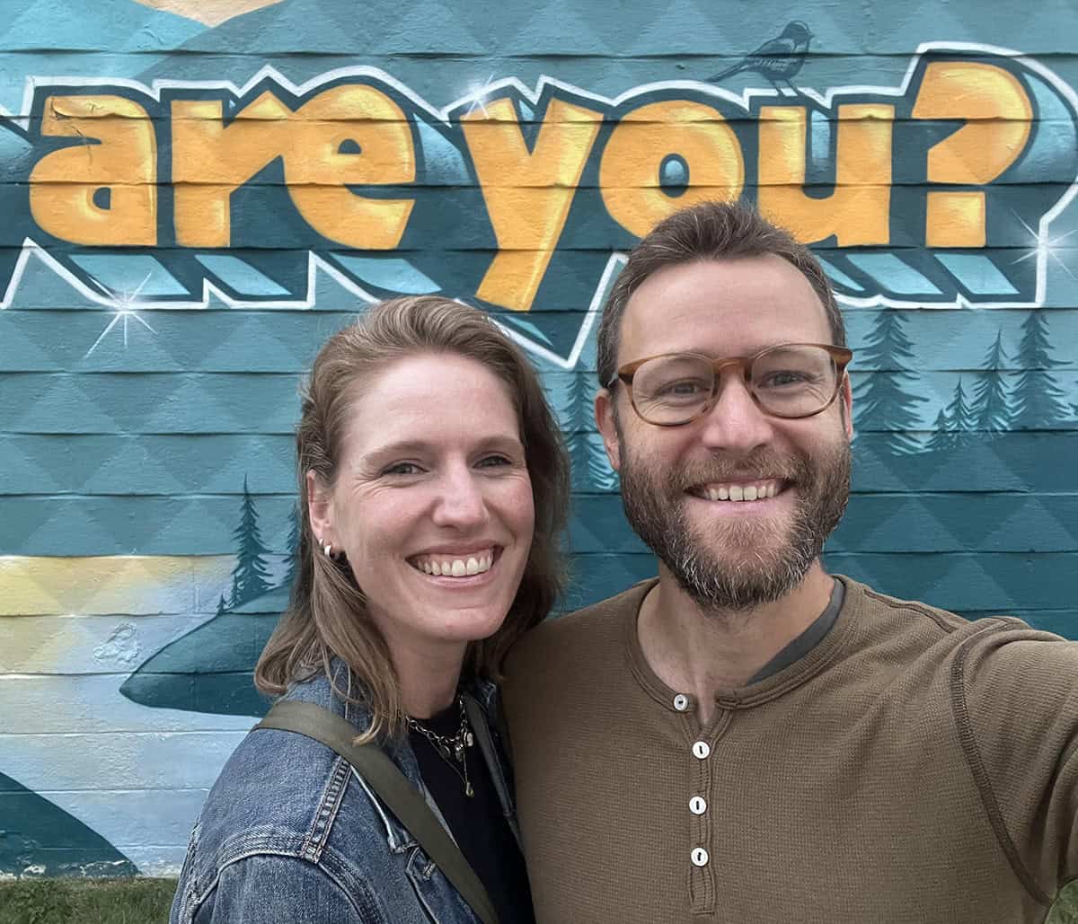 Smiling couple taking a selfie in front of a mural with 'are you?' text.